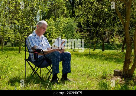 un uomo anziano che si rilassa sulla sedia da giardino leggendo un libro caldo di primavera nella contea di zala in ungheria Foto Stock