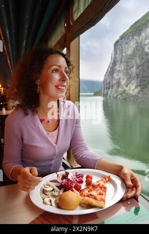 Una donna sorridente si siede a tavola e mangia davanti alla finestra sul mare Foto Stock