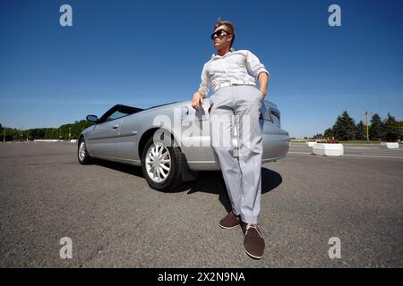 Un giovane con occhiali da sole scuri si appoggia al gomito sul bagagliaio di un'auto aperta argentata Foto Stock