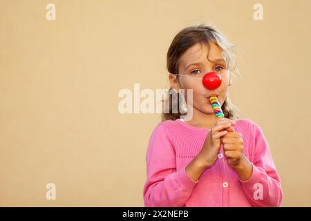 La ragazza che indossa una blusa rosa con il naso rosso clown succhia caramelle multicolore. Foto Stock