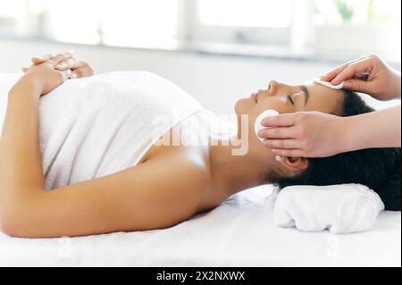 Vista laterale di una giovane donna brasiliana o ispanica ben curata, avvolta in un asciugamano, sdraiata in un salone di bellezza prima della procedura di cura della pelle del viso, la cosmetologa pulisce il viso con tamponi di cotone Foto Stock