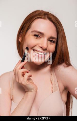 Beautiful Young Fresh Happy Redheaded Girl with A cute Smile tiene Un pennello vicino al viso in Studio su sfondo bianco. Bellezza, cura della pelle e trucco Foto Stock