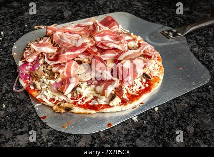 pizza irresistibile condita con prosciutto, funghi, formaggio, cipolle e ketchup, posizionato su una buccia di acciaio inossidabile su un tavolo di granito, pronto per il forno. G Foto Stock