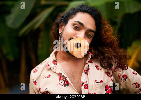 Camicia da uomo dell'Asia meridionale, occhi espressivi che tengono in bocca fiori gialli, audace femminilità, amore per se stessi in un giardino ricco. La persona rappresenta l'orgoglio LGBTQ Foto Stock