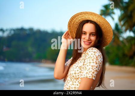 Donna sorridente in abito floreale e cappello di paglia gode di spiaggia tropicale al tramonto. Moda estiva, svago via mare, viaggiatore femminile esplora la costa, posa Foto Stock