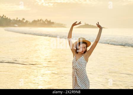 Donna in abito a pois gode del tramonto sulla spiaggia, braccia sollevate in alto. Il viaggiatore solo celebra la libertà, la calda serata estiva al mare. Felicità, relax Foto Stock