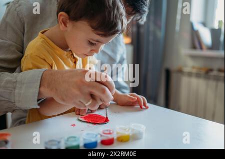 Padre che insegna al figlio a dipingere a casa uova di Pasqua in cartone con acquerelli Foto Stock