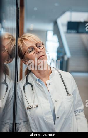 Medico appoggiato con gli occhi chiusi sul vetro in ospedale Foto Stock
