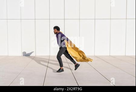 Uomo giocoso che corre di fronte a un muro bianco con indosso un mantello Foto Stock