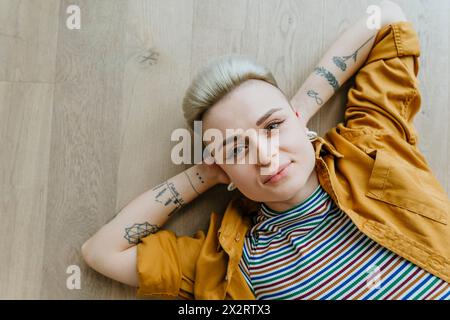Donna sorridente con tatuaggi sdraiati sul pavimento di casa Foto Stock