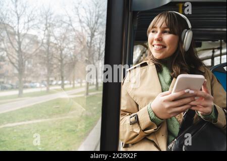 Passeggero felice che indossa le cuffie wireless che guarda dal finestrino e viaggia in autobus Foto Stock