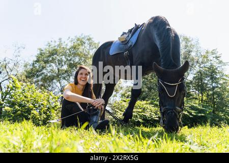 Felice istruttore seduto al pascolo del cavallo nero nelle giornate di sole Foto Stock