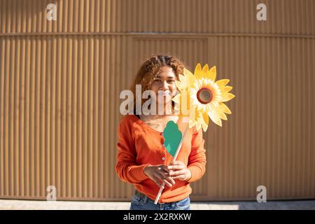 Donna sorridente che tiene un girasole artificiale davanti a una parete di metallo Foto Stock