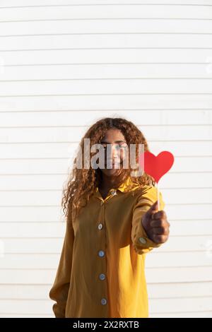 Giovane donna dai capelli ricci che dona un puntale rosso a forma di cuore davanti all'otturatore bianco Foto Stock