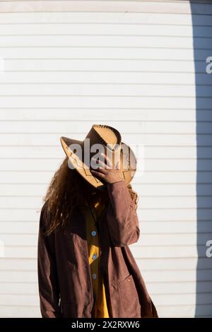 Giovane donna che indossa un cappello da cowboy e una giacca in piedi davanti a una tapparella bianca Foto Stock