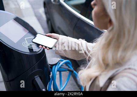 Giovane donna che paga tramite smartphone presso la stazione di ricarica per veicoli elettrici Foto Stock