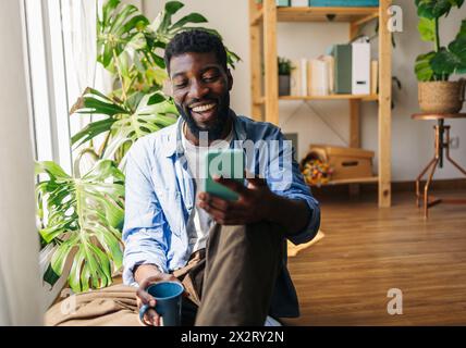 Allegra persona non binaria che usa lo smartphone e si siede con una tazza di caffè a casa Foto Stock