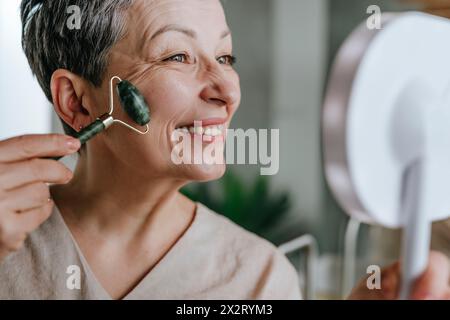 Donna felice che usa il rullo di giada sul viso a casa Foto Stock