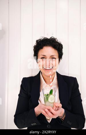 Donna d'affari felice in piedi con un bicchiere di succo di cetriolo davanti al muro Foto Stock