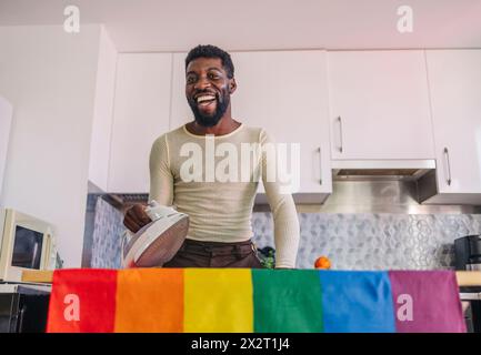 Una persona felice non-binaria che stirava la bandiera arcobaleno a casa Foto Stock