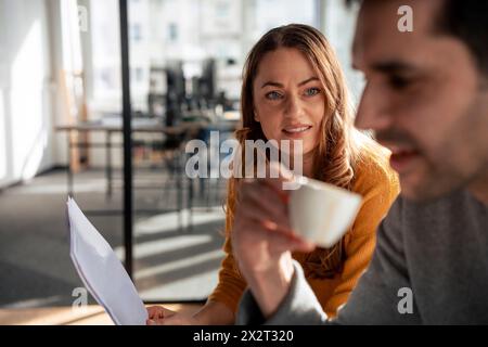 Donna d'affari sorridente che tiene un documento con un collega che prende un caffè in ufficio Foto Stock