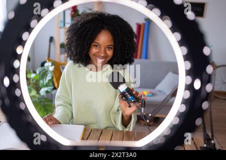 Giovane donna felice con microfono davanti alla luce anulare illuminata a casa Foto Stock