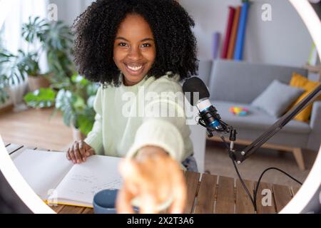 Giovane donna sorridente con microfono davanti alla luce anulare illuminata a casa Foto Stock