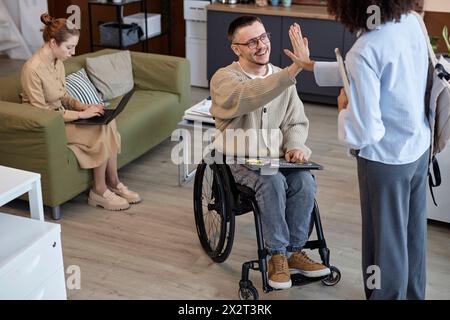 Donna d'affari che dà cinque voti a un collega disabile seduto in sedia a rotelle in ufficio Foto Stock
