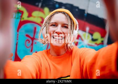 Giovane donna felice che indossa le cuffie wireless e scatta selfie davanti a una parete di graffiti Foto Stock