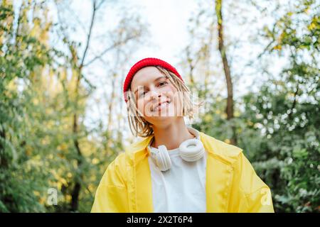 Giovane donna sorridente che indossa un cappello in maglia e un impermeabile giallo al parco Foto Stock