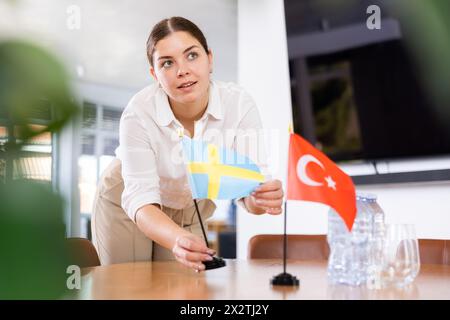 Giovane assistente donna che mette in tavola bandiere turche e svedesi per i negoziati internazionali Foto Stock