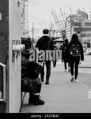 Una persona in un gilet di pelle nera indossato e cucito seduta su una panchina, in attesa dell'autobus, tenendo il suo biglietto e guardando in fondo alla strada. Foto Stock