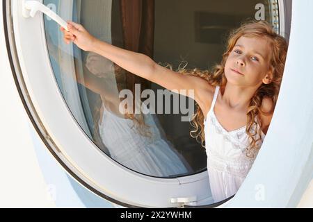 La bambina in abito bianco apre la finestra fatta come oblò Foto Stock