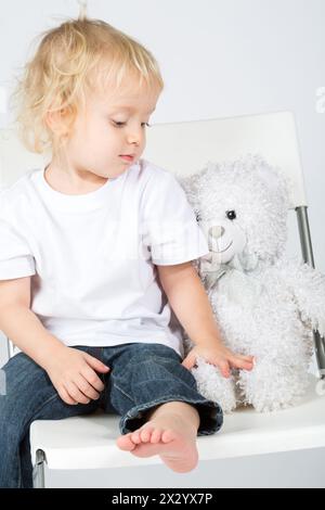 Il bambino in jeans con un orso giocattolo siede su una sedia Foto Stock
