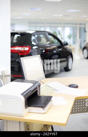 Computer, stampante e documenti su una scrivania in legno e nuovi stand per auto nell'ufficio del negozio di automobili. Profondità di campo ridotta. Messa a fuoco sul display. Foto Stock