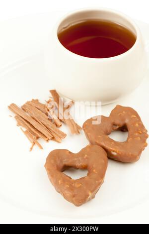 Due biscotti a forma di cuore, tazza con tè e cannella sul piatto Foto Stock