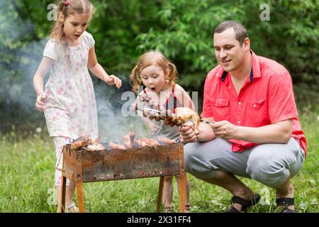 Una famiglia di tre persone che preparano barbecue sulla griglia della natura, una bambina che soffia su uno spiedino con i funghi. Foto Stock