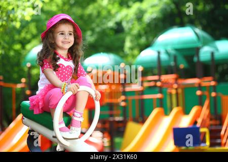 Sorridente bambina in un cappello rosa seduta su un'altalena sul parco giochi Foto Stock