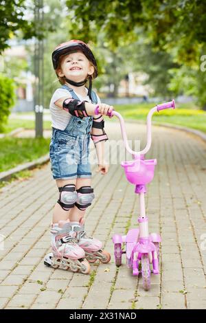 La bambina sorridente con il rullo e l'attrezzatura protettiva si trova sul passaggio pedonale del parco con in mano uno scooter a tre ruote Foto Stock
