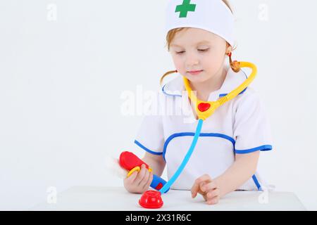 Bambina vestita da infermiera gioca con il fonendoscopio giocattolo e gli strumenti medici su sfondo bianco. Foto Stock