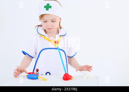 Bambina vestita da infermiera gioca con strumenti medici giocattolo su sfondo bianco. Foto Stock
