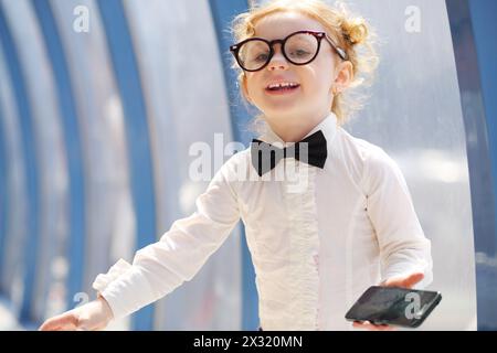 La bambina con gli occhiali tiene il cellulare nero e ride vicino alla finestra. Foto Stock