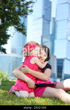 La bella ragazza con un tablet pc abbraccia la bambina sull'erba vicino ai grattacieli blu. Foto Stock