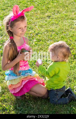 Bambina con uova di Pasqua in mano e orecchie di coniglio in testa con bambino seduto sull'erba Foto Stock