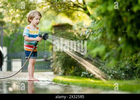 Un bambino carino che gioca con il tubo da giardino nelle calde giornate estive. Bambino che gioca con l'acqua in estate. Divertimento attivo con bambini piccoli. Foto Stock