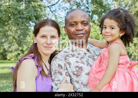 Ritratto di madre bianca e padre nero, che tiene una figlia piccola sulle mani, in piedi nel parco estivo Foto Stock