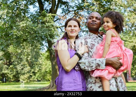 Ritratto di madre bianca e padre africano, che tiene una figlia piccola sulle mani, in piedi nel parco estivo Foto Stock