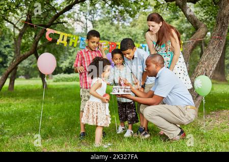 Il padre accende la candela sulla torta di compleanno con accendino, la famiglia di cinque persone la guarda, il cartello di buon compleanno dietro le spalle Foto Stock