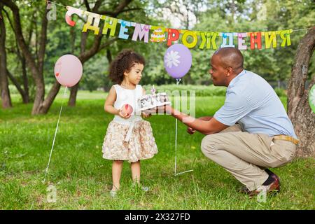 La bambina si trova nel parco, tenendo in mano la grande mela rossa, il padre le regala la torta di compleanno, il cartello di buon compleanno alle spalle Foto Stock