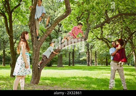 La famiglia di sei persone si riposa nel parco, i ragazzi si arrampicano sull'albero, il padre gioca con la figlia piccola, la madre si fa da parte Foto Stock